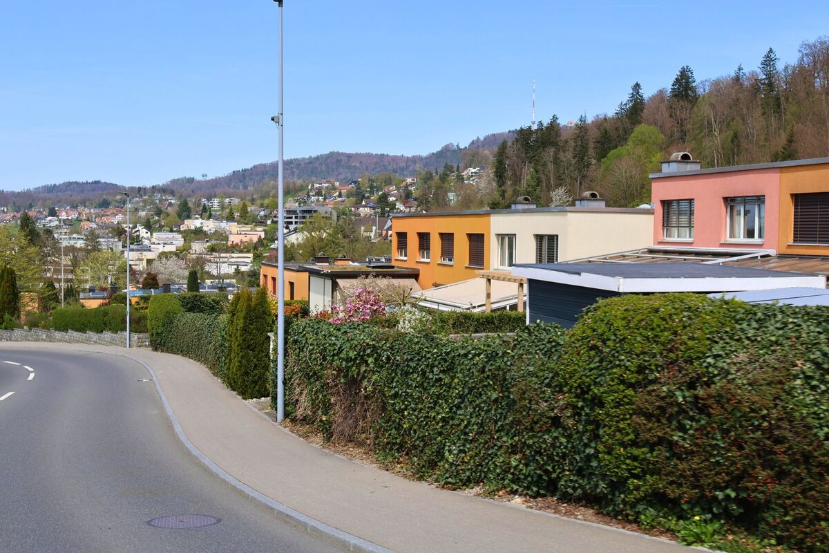  Moderne Einfamilienhäuser mit farbigen Fassaden reihen sich entlang einer Wohnstrasse in Bonstetten im PLZ-Gebiet 8906 im Kanton Zürich, mit Blick auf das Dorf und bewaldete Hügel.