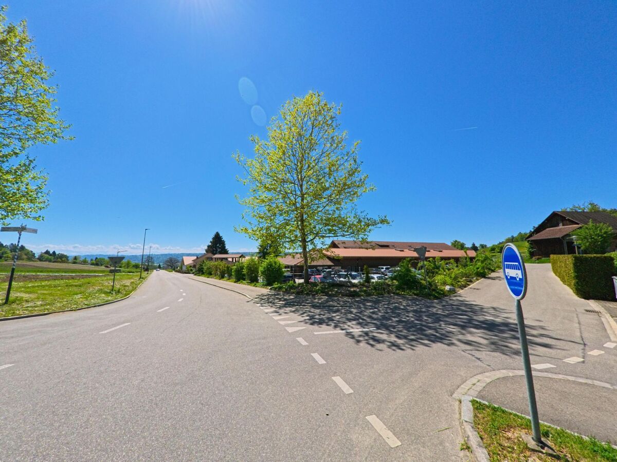 Eine weite Kreuzung mit Buswendeschleife liegt am sonnigen Dorfrand von Boppelsen im PLZ-Gebiet 8113 im Kanton Zürich, mit Blick auf Felder und Hügel in der Ferne.