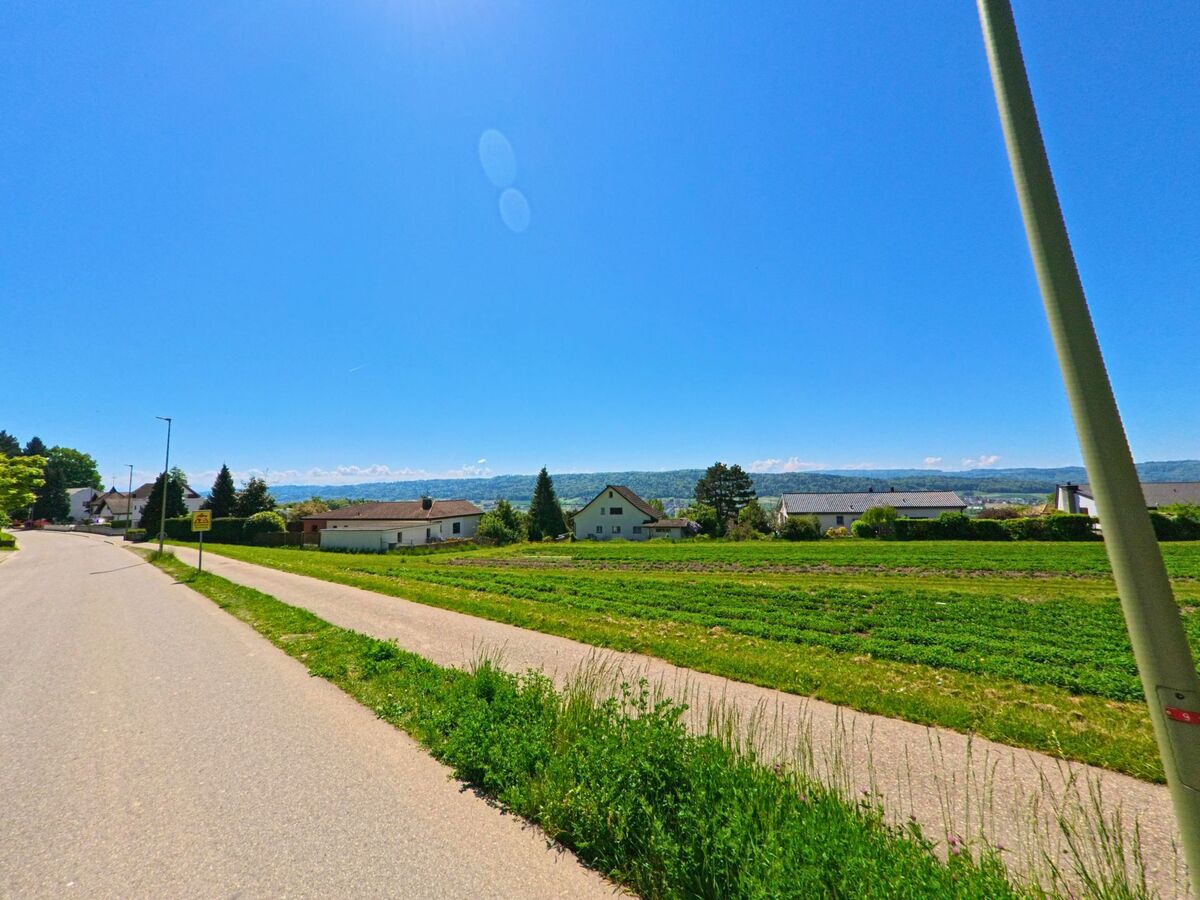 Am Rand von Boppelsen im PLZ-Gebiet 8113 im Kanton Zürich verlaufen Strasse und Feldweg parallel zwischen gepflegten Gärten und einer weiten Aussicht auf die bewaldeten Hügel.