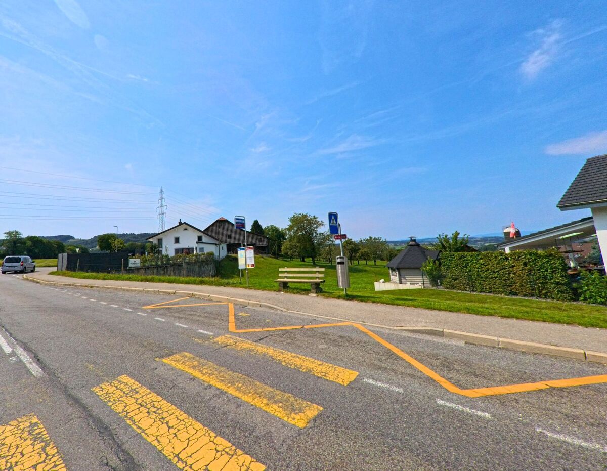 Eine Bushaltestelle mit Sitzbank und Strassenschild befindet sich am Ortsrand von Bossonnens (1615, Kanton Freiburg) mit Blick auf Wiesen und Hauser.