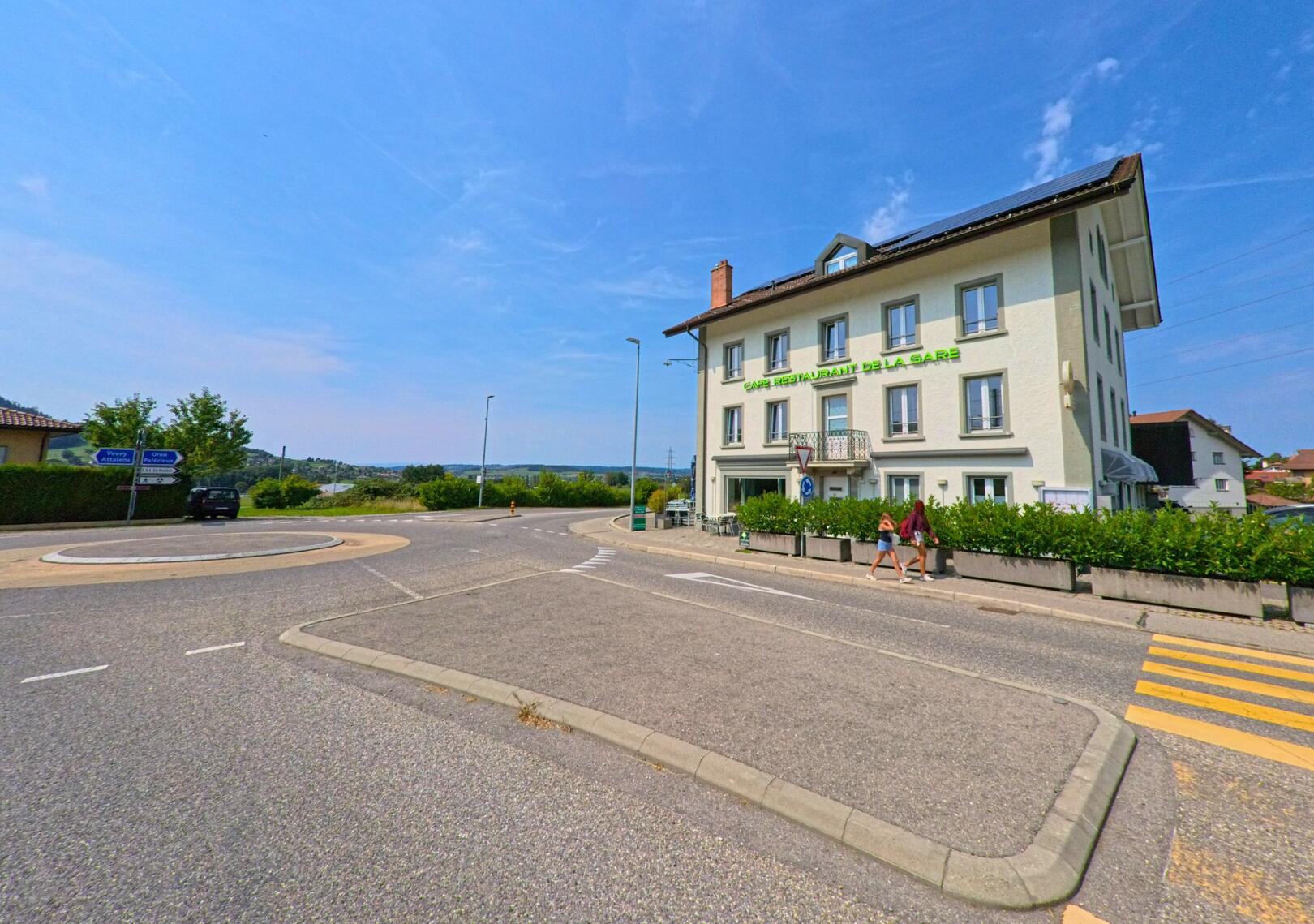 An der Strasse von Bossonnens (1615, Kanton Freiburg) steht das Café Restaurant de la Gare neben einem kleinen Kreisel, während zwei Fussgaenger am Gehweg vorbeigehen.