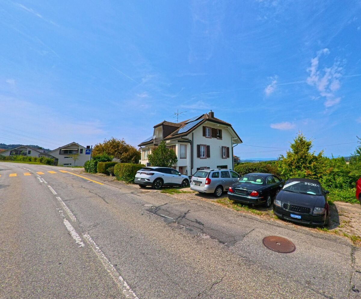 Ein Einfamilienhaus mit geparkten Autos davor befindet sich an einer Nebenstrasse in Bossonnens (1615, Kanton Freiburg) nahe einem Fussgaengerstreifen.