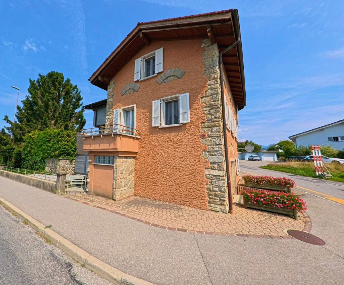 Ein Haus mit orangefarbener Fassade, weissen Fensterlaeden und Blumenschmuck steht an einer Strasse in Bossonnens (1615, Kanton Freiburg).