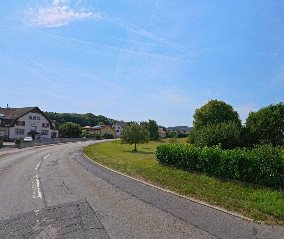 Eine leicht gekruemmte Strasse fuehrt durch ein Wohnquartier von Bossonnens (1615, Kanton Freiburg), links und rechts von Wiesen und Baeumen umgeben.
