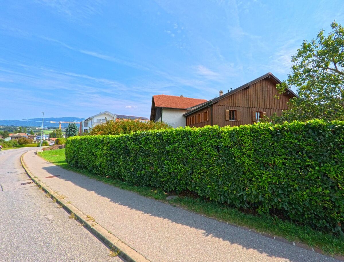 Hinter einer dichten gruene Hecke steht ein traditionelles Wohnhaus mit Holzfassade und rotem Dach in Bossonnens (1615, Kanton Freiburg).