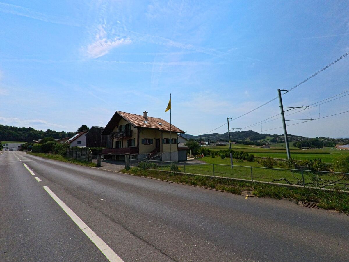 Ein Wohnhaus mit Balkon und gelber Fahne steht an einer Hauptstrasse in Bossonnens (1615, Kanton Freiburg), rechts verlaufen Eisenbahngleise durch die Landschaft.
