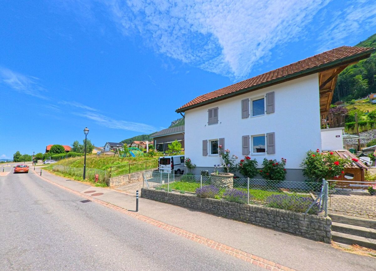 In Botterens (1652, Kanton Freiburg) steht ein weisses Einfamilienhaus mit gepflegtem Vorgarten und Blumen entlang einer leicht abfallenden Strasse.