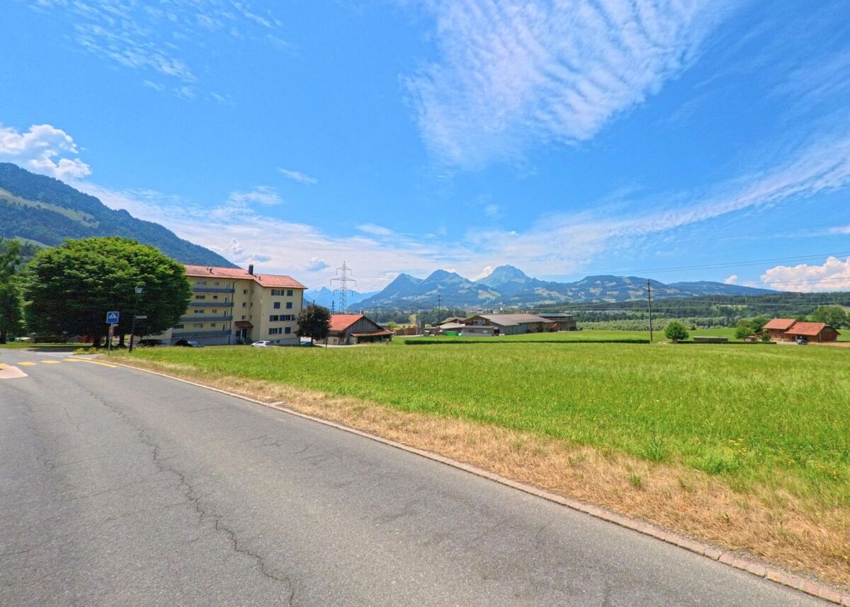 In Botterens (1652, Kanton Freiburg) verläuft eine Landstrasse mit Fussgängerstreifen durch eine offene Wiesenlandschaft, dahinter erstreckt sich ein Alpenpanorama.