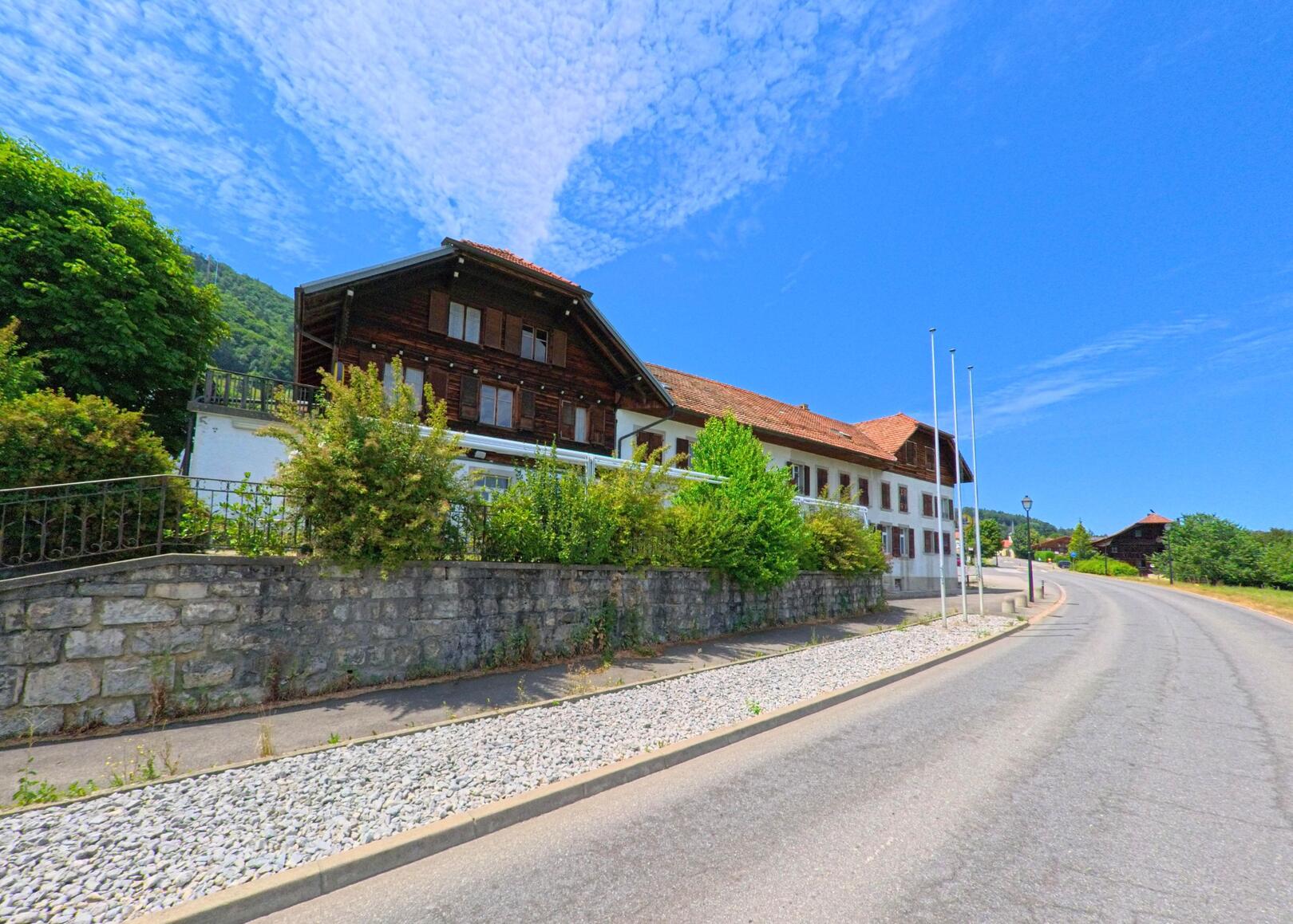 In Botterens (1652, Kanton Freiburg) säumen traditionelle Gebäude mit Holzfassaden und Ziegeldächern eine leicht ansteigende Strasse am Rand eines bewaldeten Hangs.