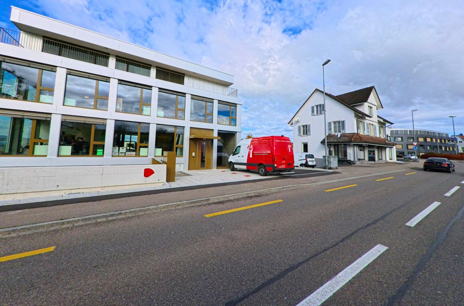 An einer Strasse in Bottighofen (8598, Kanton Thurgau) stehen moderne und traditionelle Gebaeude nebeneinander, waehrend parkierte Fahrzeuge das Ortsbild praegen.