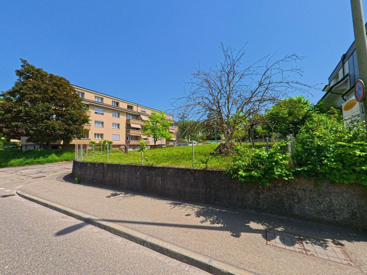 In Bottmingen (4103, Kanton Basel-Landschaft) liegt ein Mehrfamilienhaus mit Balkonen gegenüber einem leicht erhöhten Grundstück mit Wiese und einzelnen Bäumen.