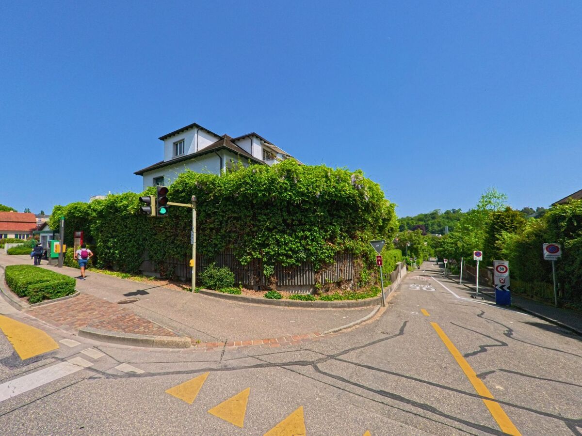 In Bottmingen (4103, Kanton Basel-Landschaft) beginnt hinter einer Ampel ein verkehrsberuhigter Strassenabschnitt mit dichter Begrünung und Fussgängerzone.