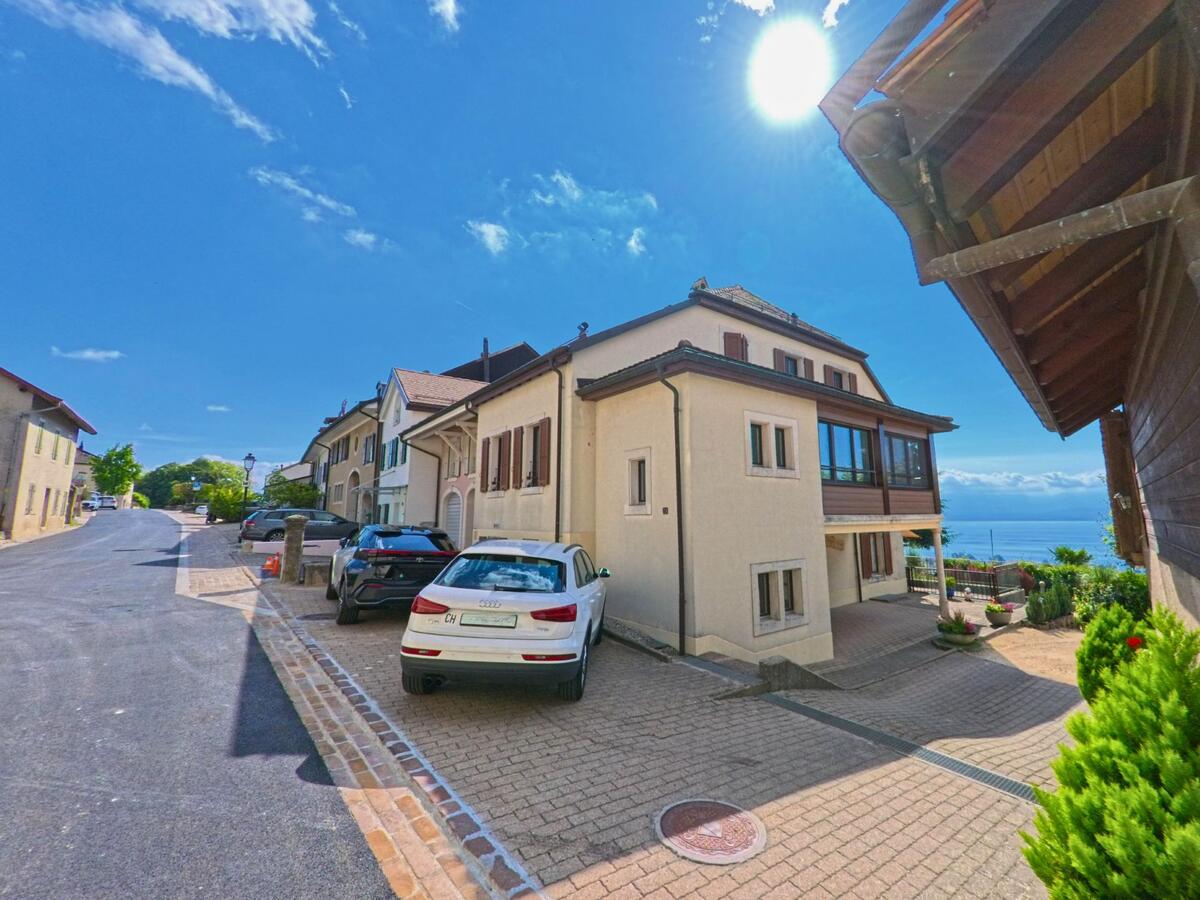 Eine sonnige Strasse in Bougy-Villars (1172, Kanton Waadt) fuehrt an Wohnhaeusern mit Blick auf den Genfersee vorbei, waehrend Autos am Strassenrand parken.