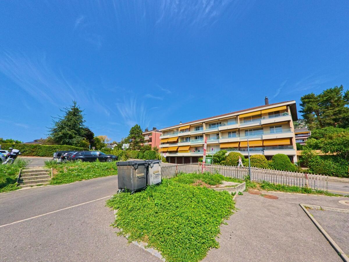 Ein Wohnblock mit gelben Markisen in Bourg-en-Lavaux (1096, Kanton Waadt) steht an einer kleinen Strasse, wo ein Parkplatz und Müllcontainer sichtbar sind.