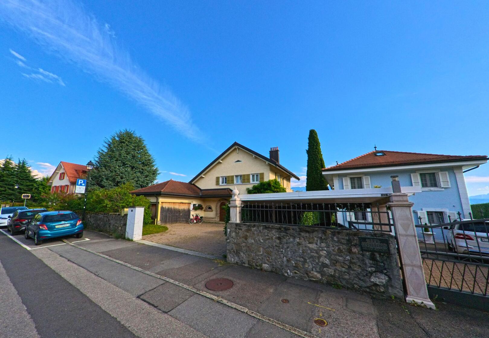 Ein Wohnhaus mit gepflegtem Vorhof in Bourg-en-Lavaux (1096, Kanton Waadt) liegt an einer ruhigen Strasse mit geparkten Autos und Bäumen im Hintergrund.