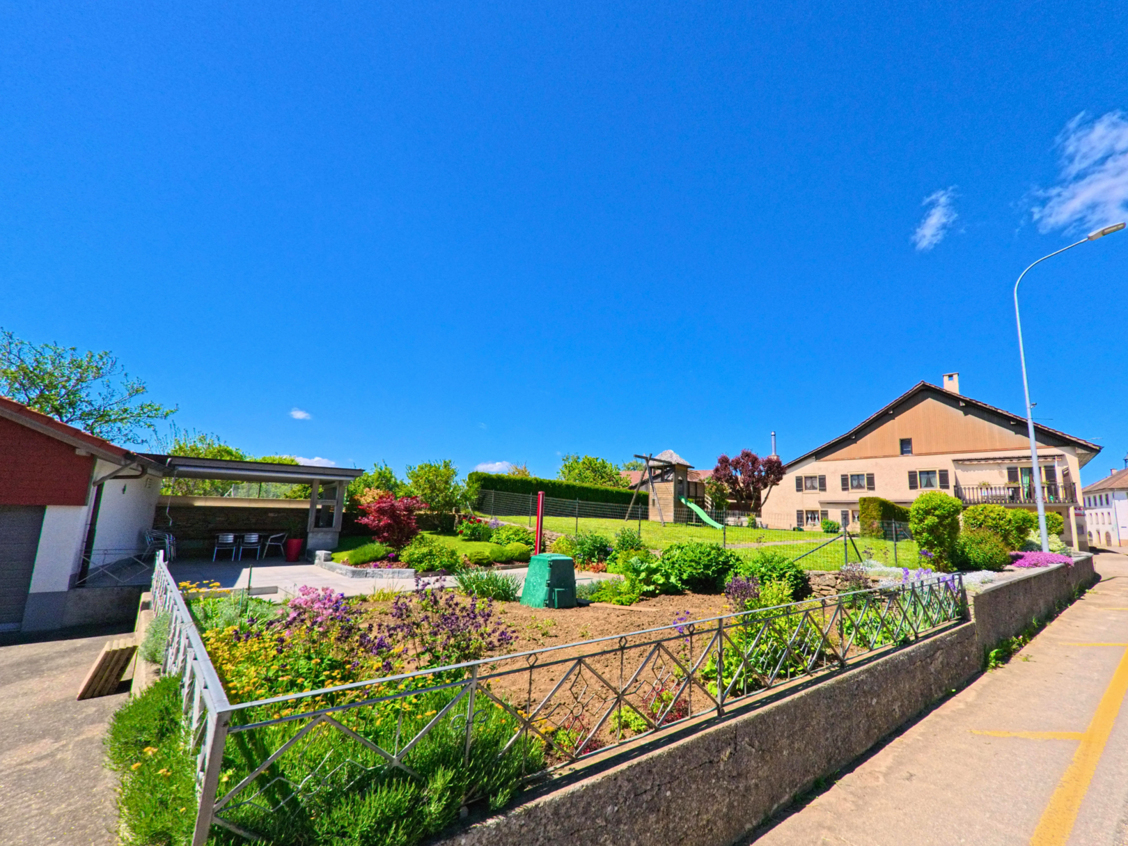In Bourrignon (2803, Kanton Jura) zeigt sich ein gepflegter Vorgarten mit Blumenbeet und Sitzplatz neben einer Wohnstrasse bei strahlend blauem Himmel.