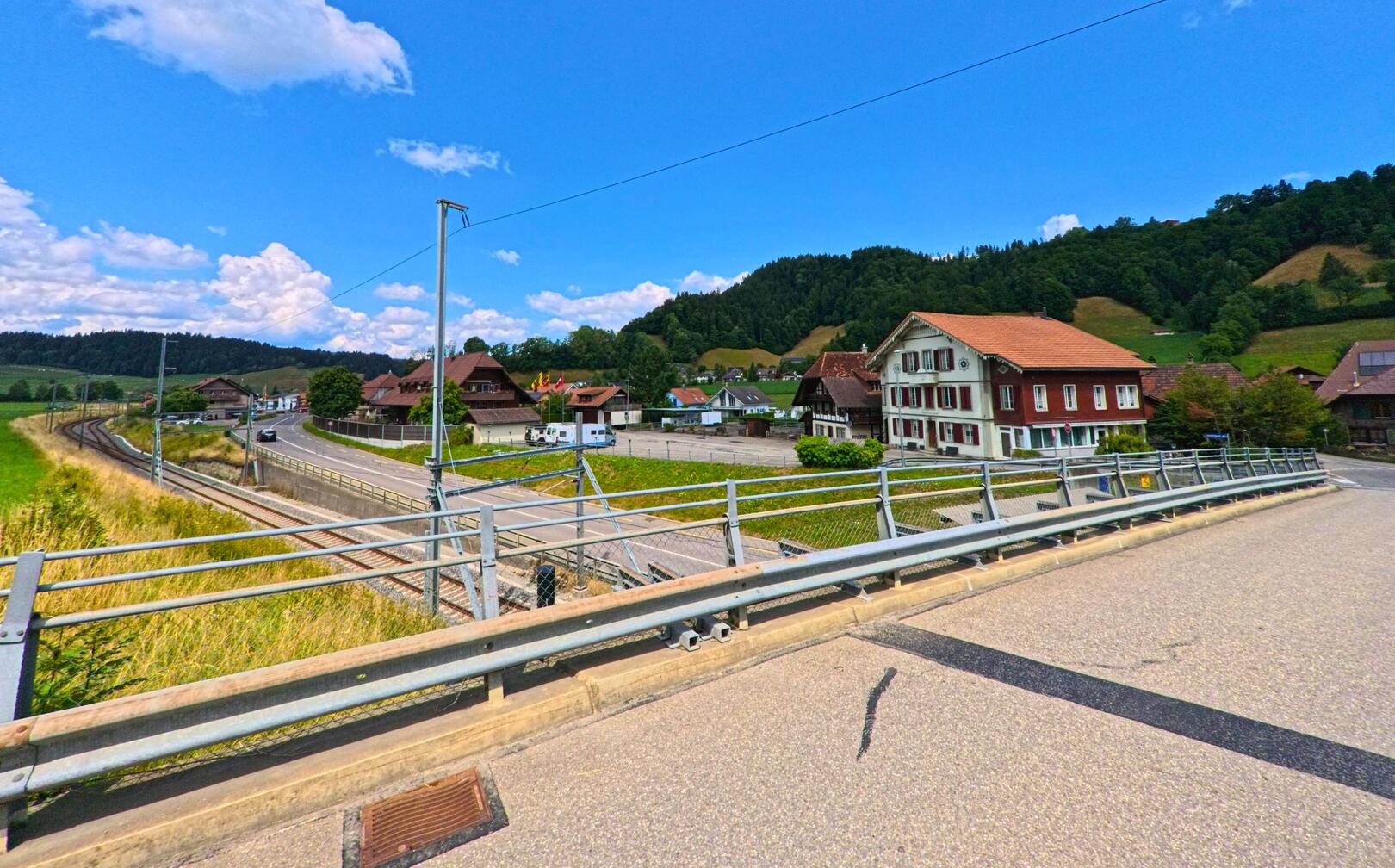 Von einer Brücke in Bowil (PLZ 3533, Kanton Bern) blickt man auf Gleise, ein markantes Haus mit roter Fassade und die umliegenden Hügel des Emmentals.
