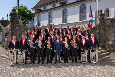 Vor der Kirche in Knutwil (6212, Kanton Luzern) steht die Brass Band Feldmusik Knutwil in festlicher Uniform mit ihren Blechblaesern zu einem Gruppenfoto auf den Steinstufen.