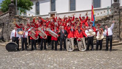 Mitglieder der Brass Band Musikgesellschaft Geiss jubeln in roter Uniform mit Instrumenten auf einer Treppe vor der Kirche in Menznau im Kanton Luzern.