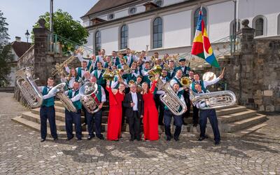 Gruppenfoto der Brass Band Musikgesellschaft Hasle auf einer Treppe vor der Kirche in Hasle, 6166, Kanton Luzern, mit zahlreichen Musikerinnen und Musikern, grossen Blasinstrumenten, zwei Personen in roten Kleidern und einer farbigen Fahne.