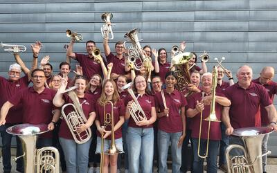 Vor einer Metallfassade in Birmenstorf (5413, Kanton Aargau) posiert die Brass Band Musikverein Birmenstorf in einheitlicher Vereinskleidung mit zahlreichen Blechblasinstrumenten für ein gemeinsames Gruppenbild.