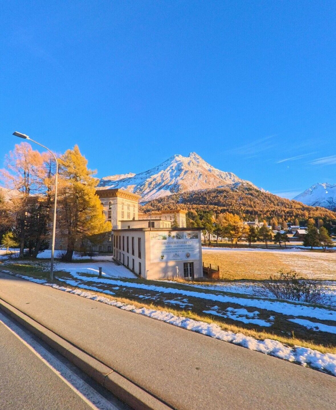 Am Rand einer offenen Wiesenfläche in Bregaglia (7516, Kanton Graubünden) steht ein markantes Gebäude vor schneebedeckten Bergflanken und herbstlich gefärbten Bäumen.