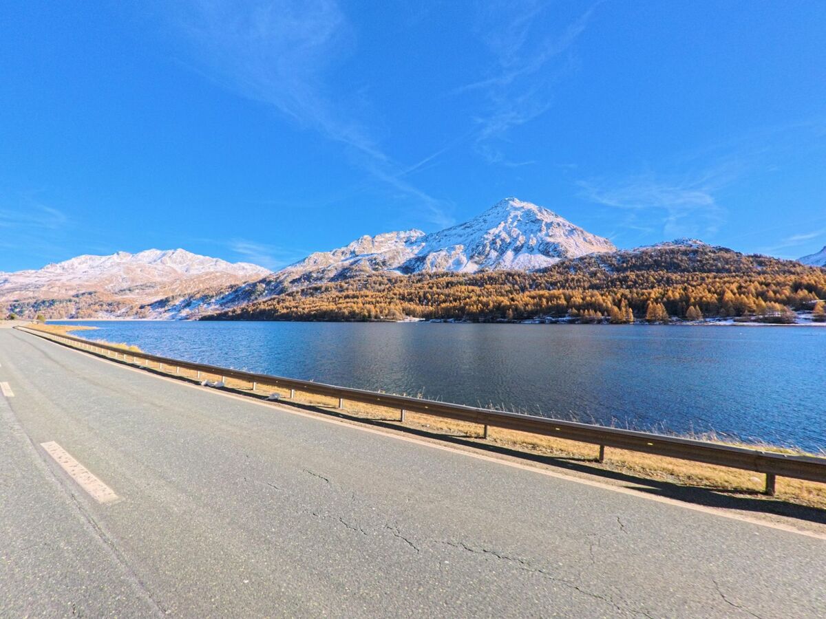 Entlang einer Uferstrasse in Bregaglia (7516, Kanton Graubünden) liegt ein See vor schneebedeckten Berggipfeln und herbstlich gefärbten Wäldern.