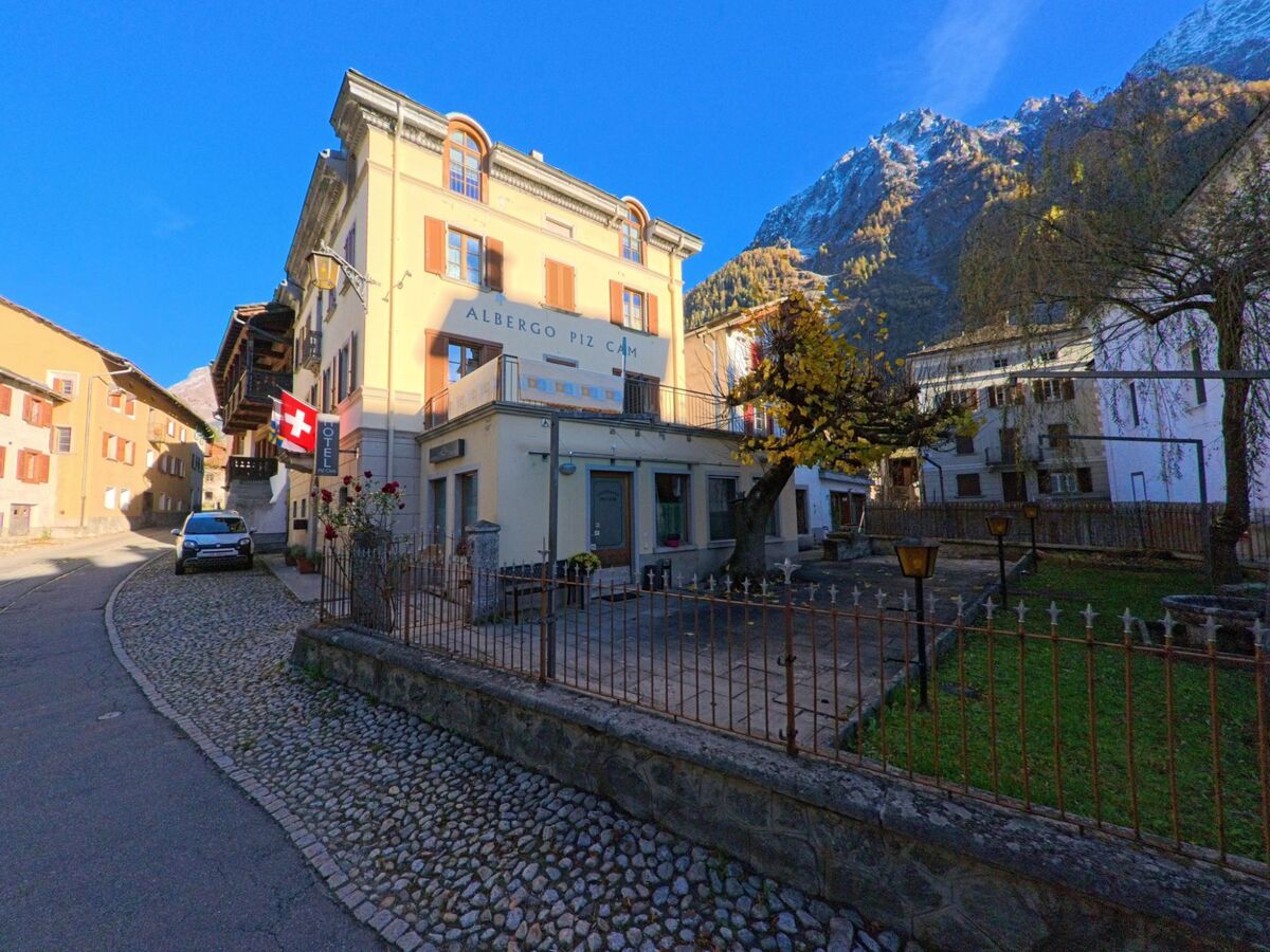 An einer gepflasterten Gasse in Bregaglia (7603, Kanton Graubünden) steht ein historisches Gebäude mit Gartenanlage vor steilen, herbstlich gefärbten Berghängen.