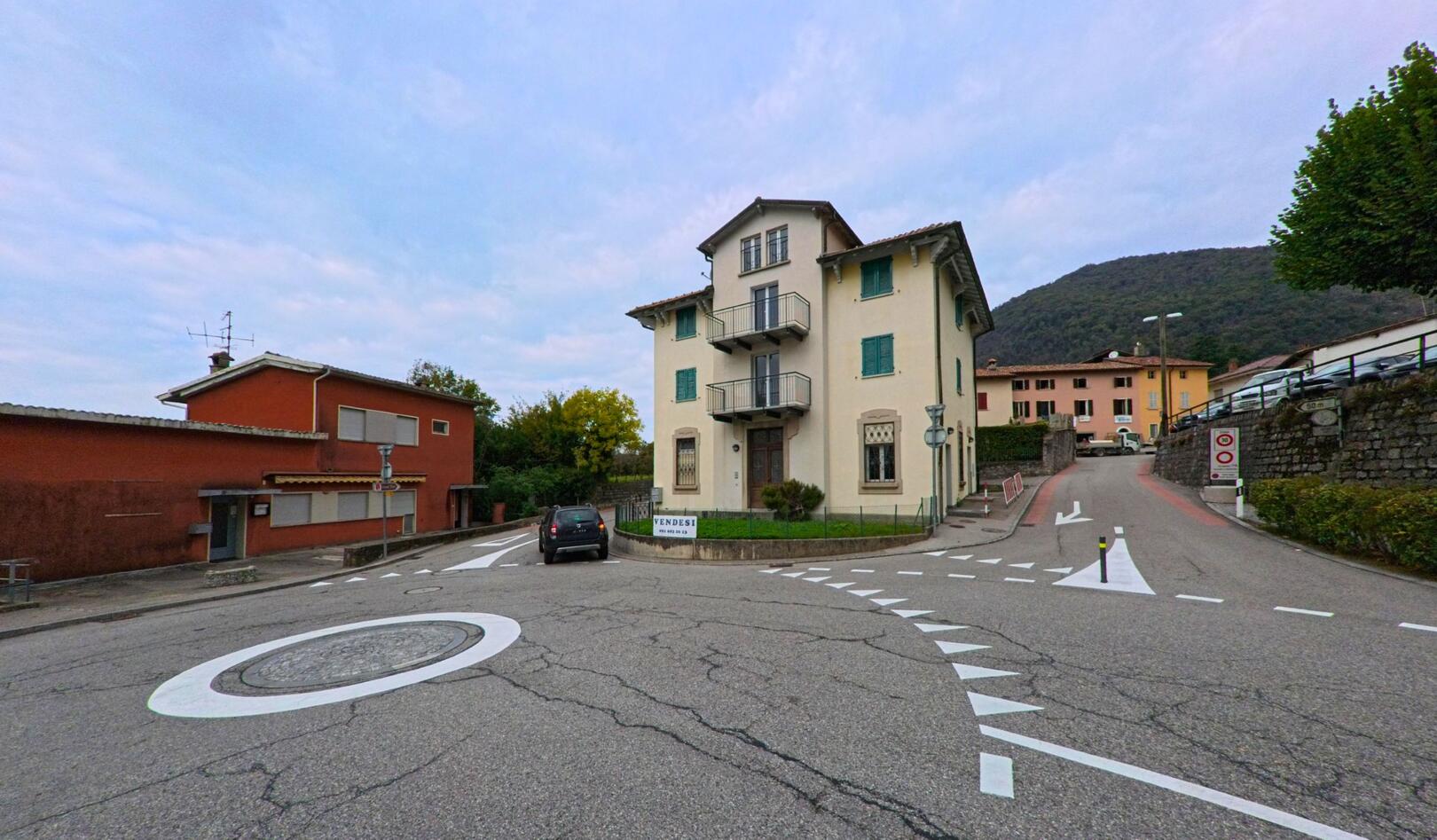 An einer kleinen Kreuzung in Breggia (6835, Kanton Tessin) steht ein helles Wohnhaus mit grünen Fensterläden vor bewaldeten Hügeln.
