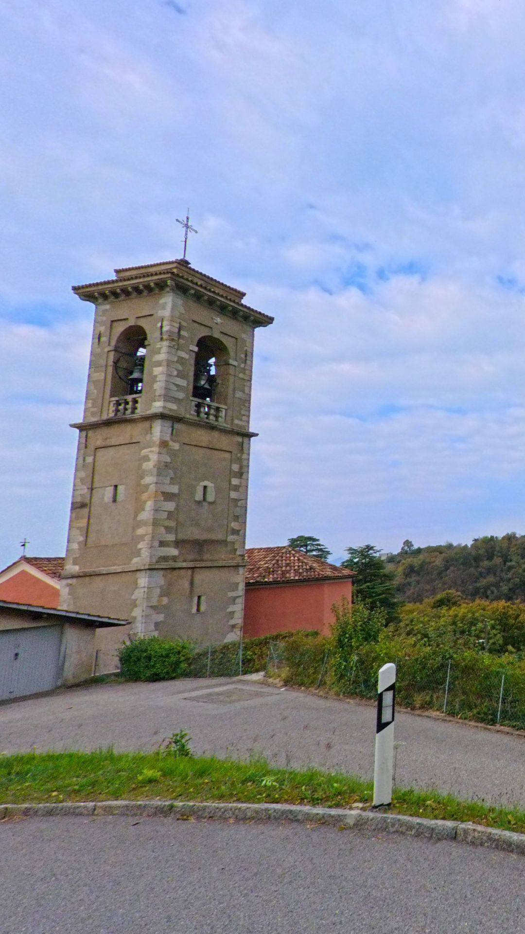 Der Kirchturm von Breggia (6835, Kanton Tessin) steht mit seinem hellen Mauerwerk und dem Kreuz auf dem Dach vor einem leicht bewölkten Himmel.