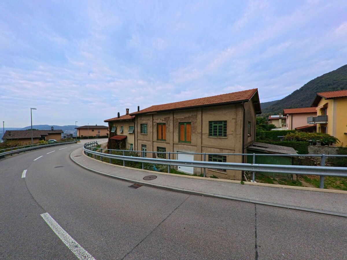 Eine leicht geschwungene Strasse in Breggia (6835, Kanton Tessin) fuehrt an einem alten Wohnhaus mit grünen Fensterrahmen und roten Ziegeldach vorbei.