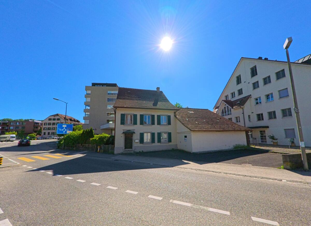 In Breitenbach (4226, Kanton Solothurn) steht ein traditionelles Wohnhaus mit gruenen Fensterlaeden im Kontrast zu den modernen Gebaeuden der Umgebung.