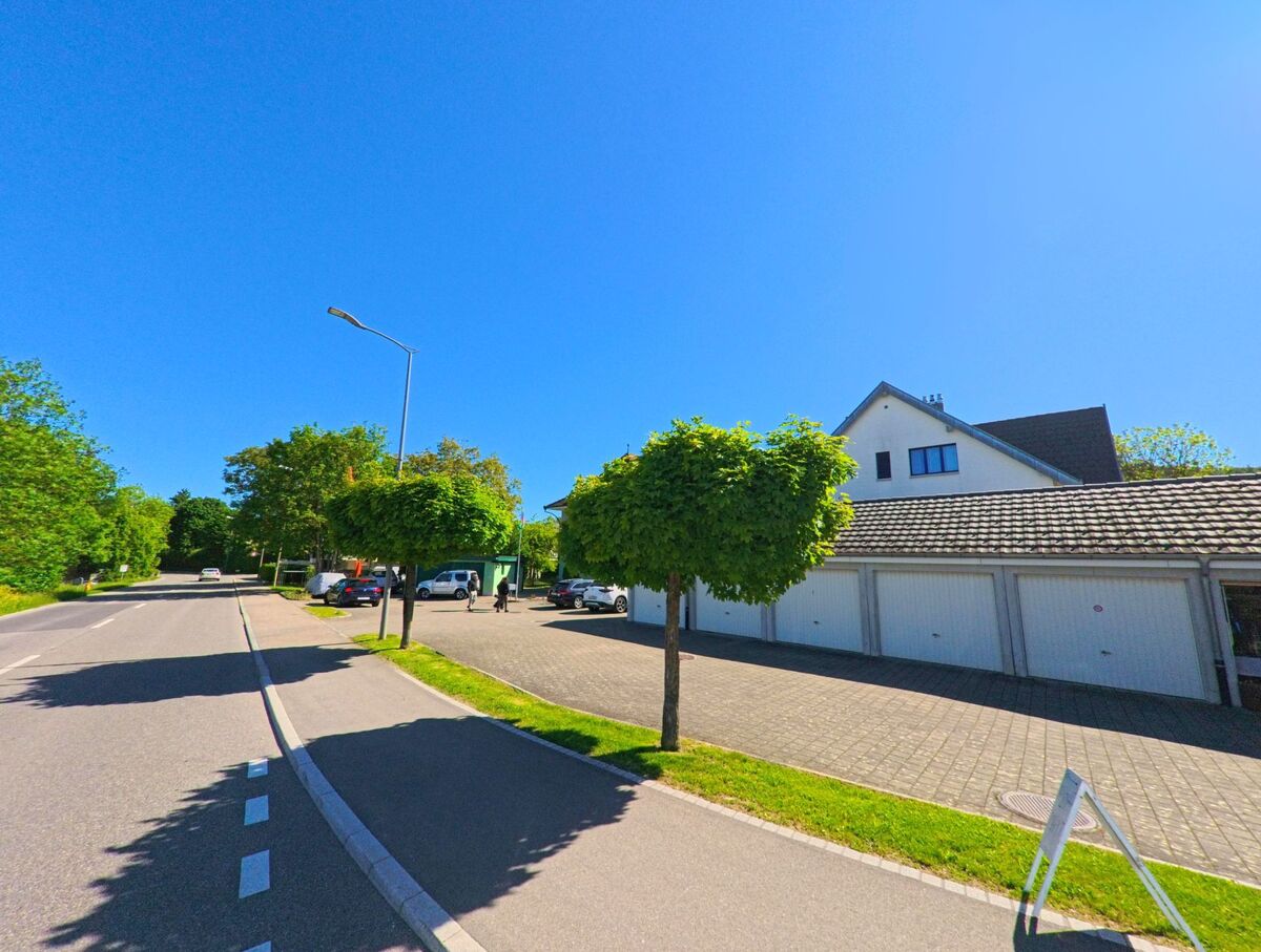 In Breitenbach (4226, Kanton Solothurn) verlaeuft eine ruhige Strasse mit Gehweg, gesaeumt von Garagen, Baeumen und einem kleinen Parkplatz.