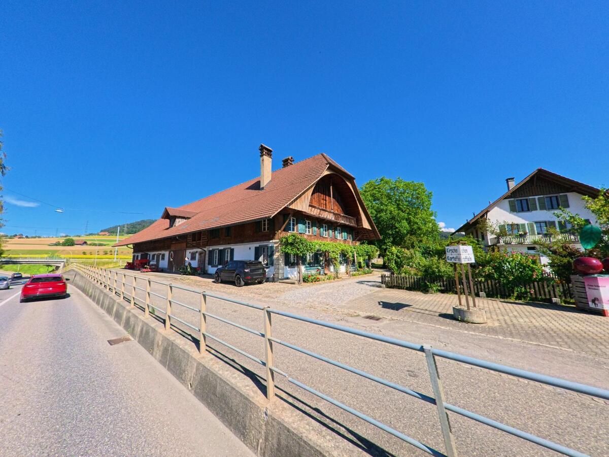 Ein traditionelles Bauernhaus mit grossem Dach steht in Brenzikofen (3671, Kanton Bern) an einer Durchfahrtsstrasse, umgeben von Feldern und weiteren Wohngebäuden.