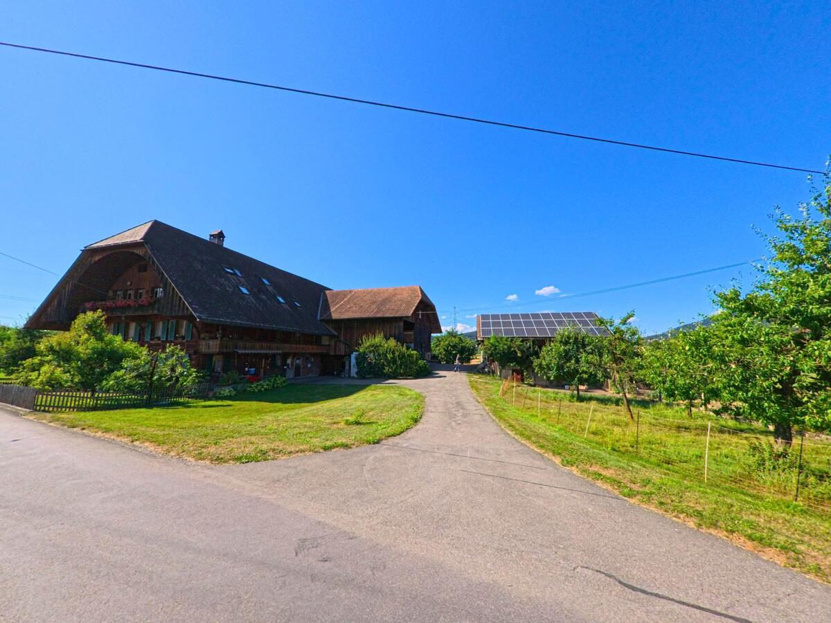 Ein landwirtschaftliches Anwesen in Brenzikofen (3671, Kanton Bern) mit traditionellem Bauernhaus und einem Nebengebäude mit Solarpanels liegt an einem geteerten Weg zwischen Obstbäumen.