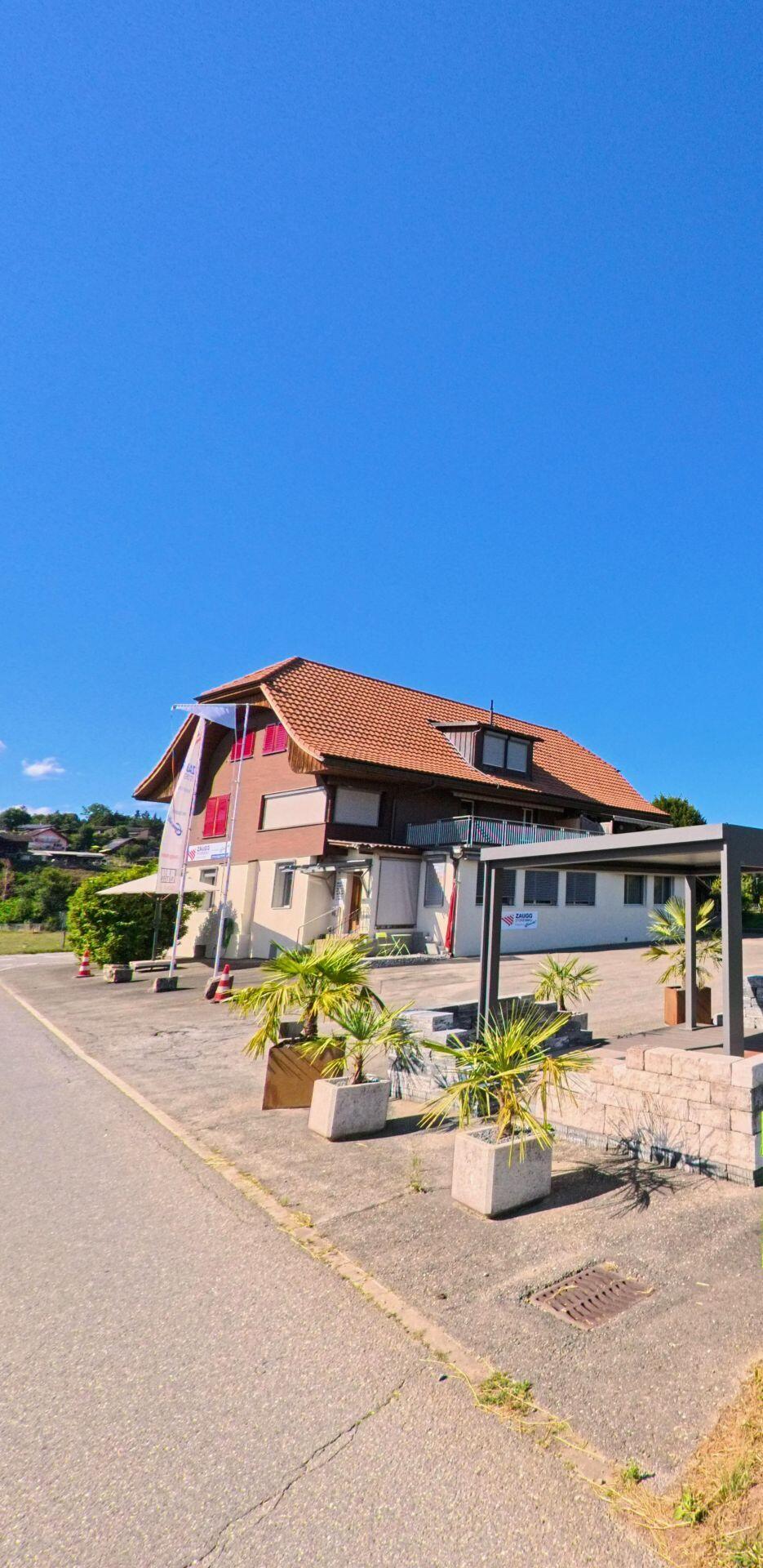 Vor einem Gebäude mit rotem Dach in Brenzikofen (3671, Kanton Bern) stehen mehrere Pflanzkübel mit Palmen entlang einer Einfahrt.