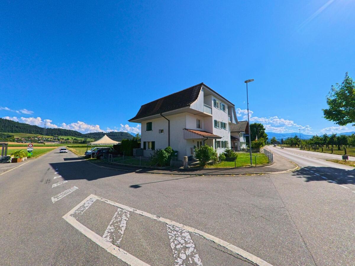 In Brenzikofen (3671, Kanton Bern) treffen zwei Strassen vor einem hellen Wohnhaus aufeinander, während im Hintergrund Felder und Hügel unter klarem Himmel liegen.