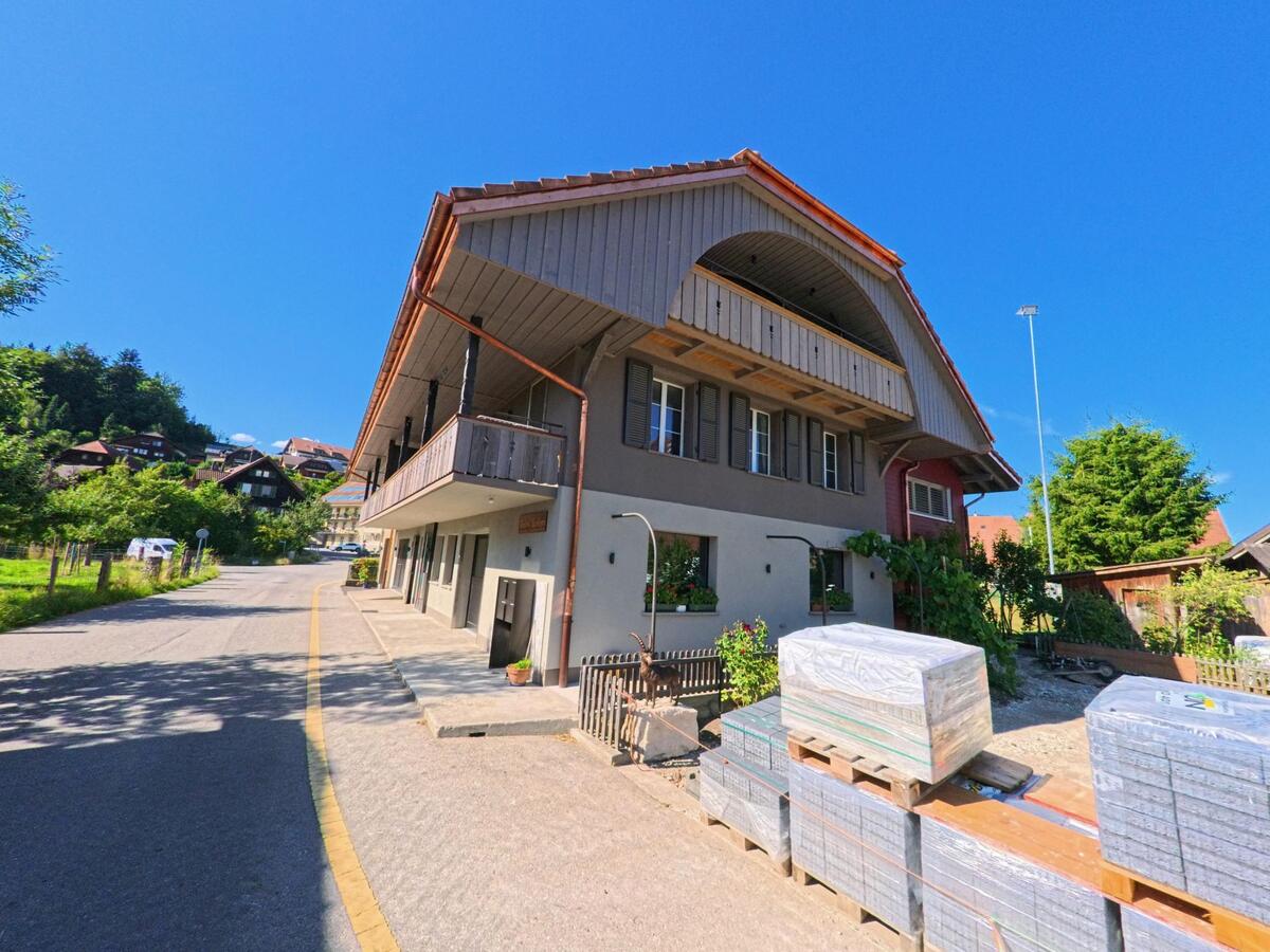 In Brenzikofen (3671, Kanton Bern) steht ein traditionelles Haus mit grossem Balkon an einer leicht ansteigenden Strasse, während gestapelte Baumaterialien im Vordergrund liegen.