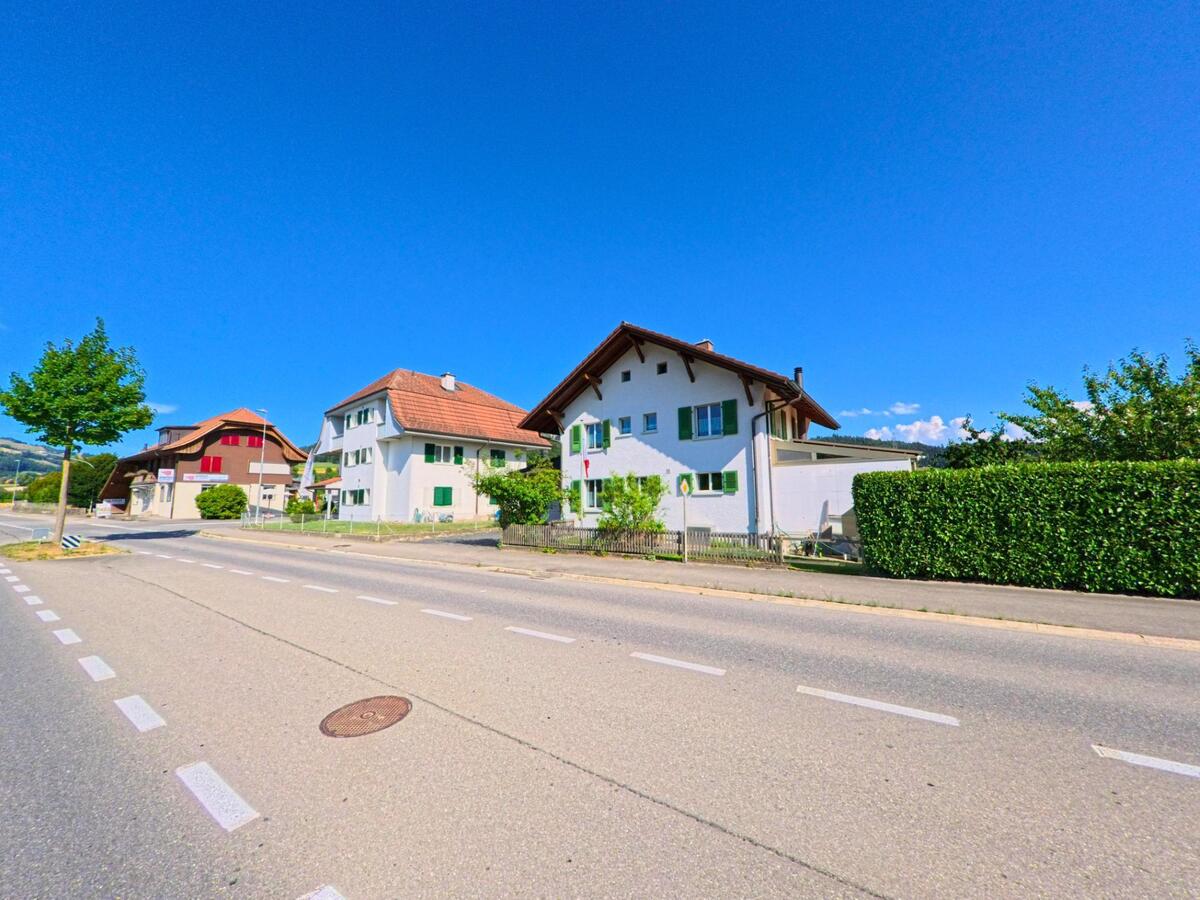 Mehrere Wohnhäuser mit farbigen Fensterläden stehen in Brenzikofen (3671, Kanton Bern) entlang einer breiten Strasse unter wolkenlosem Himmel.