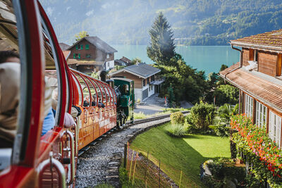 In Brienz (3855, Kanton Bern) faehrt die rote Zahnradbahn der Brienz Rothorn Bahn AG an Hausern vorbei mit Blick auf den See und die umliegenden Berge.