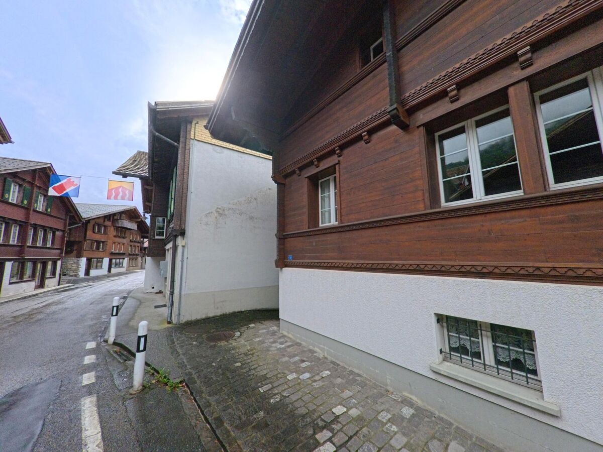 Zwischen traditionellen Holzhäusern mit verzierten Fassaden verläuft in Brienz (3855, Kanton Bern) eine enge, gepflasterte Gasse mit aufgehängten Gemeindefahnen.