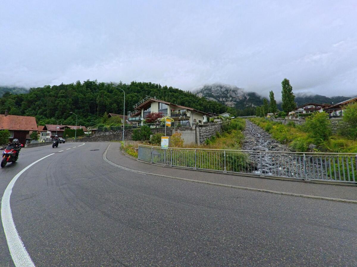 In Brienz (3855, Kanton Bern) fliesst ein steiniger Bachlauf zwischen Wohnhäusern und Bäumen hinab, während sich eine kurvige Strasse mit Motorradfahrern daran vorbeischlängelt.