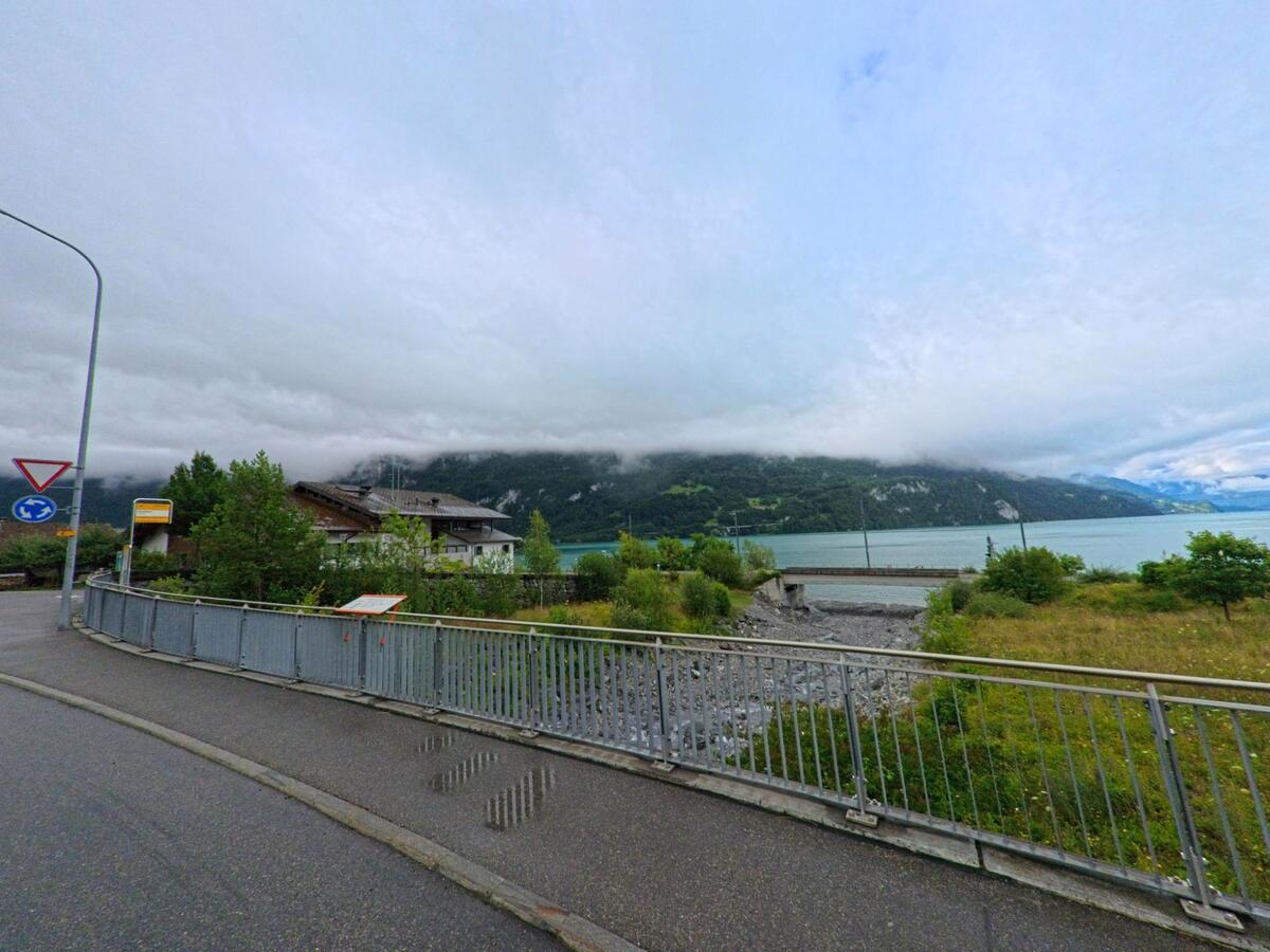 In Brienz (3855, Kanton Bern) führt ein Gehweg mit Geländer an einer Bushaltestelle und einem Bachbett entlang, mit Blick auf den wolkenverhangenen Brienzersee.