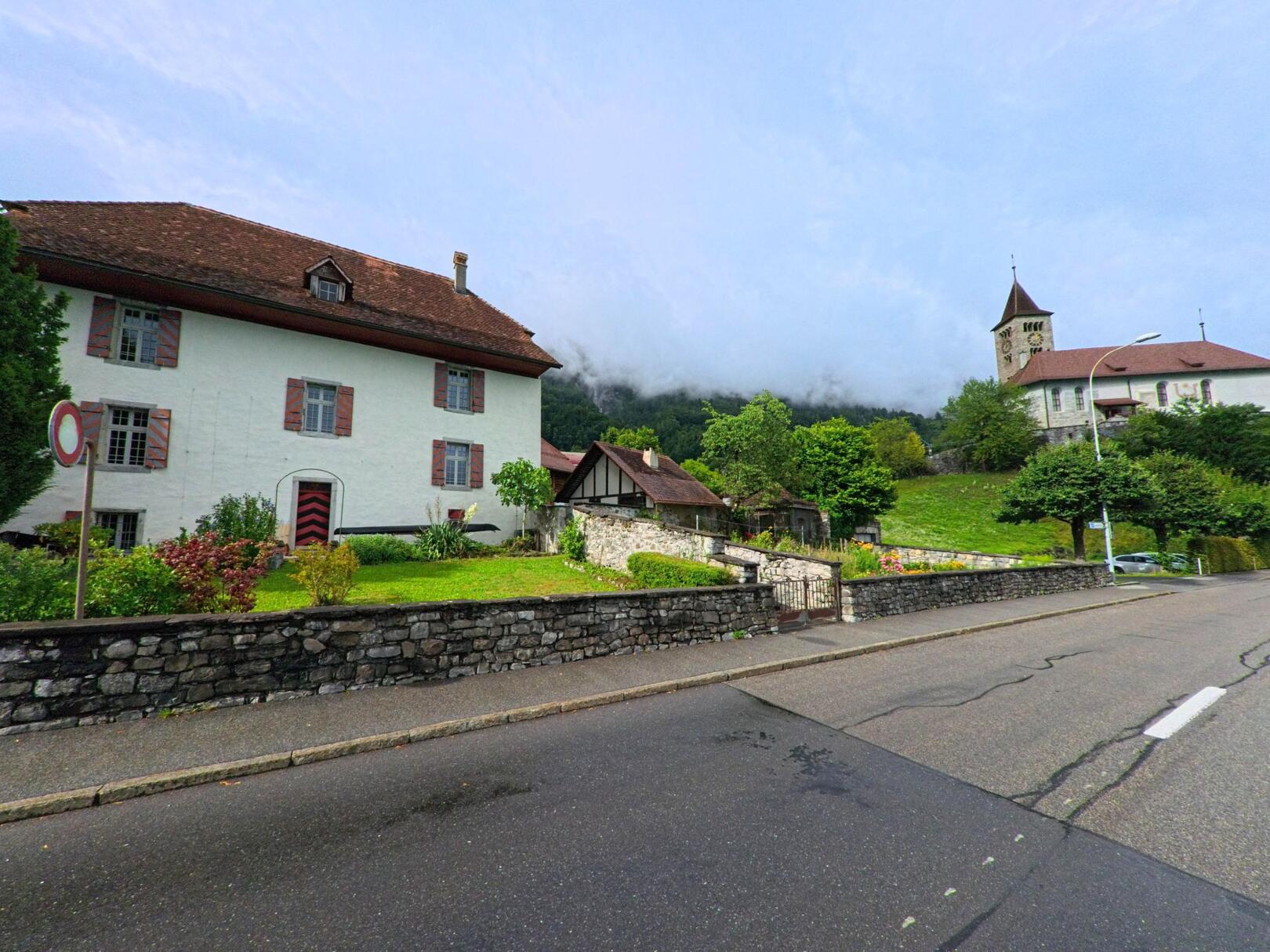 In Brienz (3855, Kanton Bern) erhebt sich neben einer historischen Kirche ein hell verputztes Wohnhaus mit rot-weiss gestrichenen Fensterlaeden und gepflegtem Vorgarten.