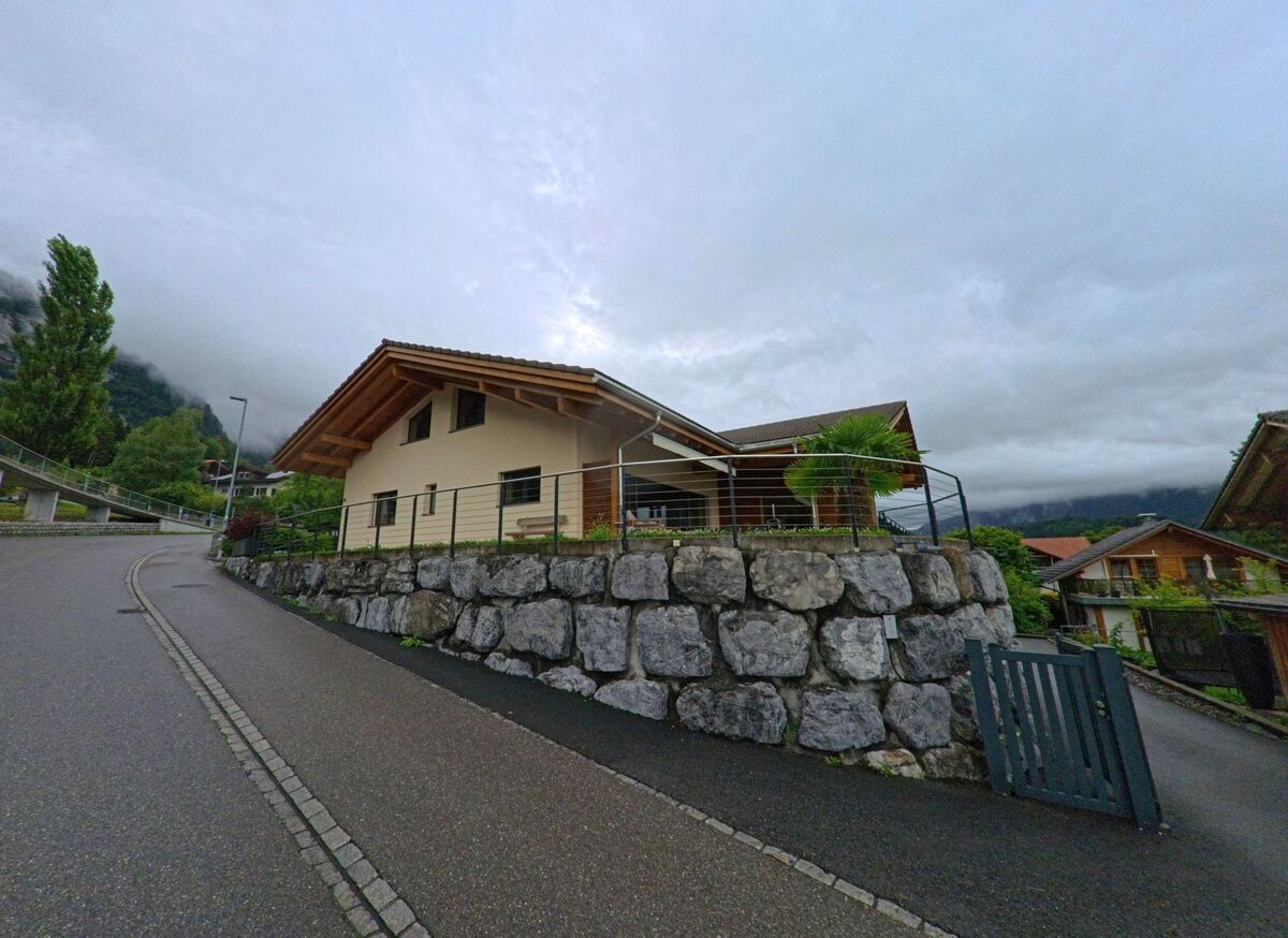 Ein Einfamilienhaus mit Flachdach in Brienz (3855, Kanton Bern) steht auf einer mit grossen Natursteinen befestigten Terrasse an einer leicht ansteigenden Strasse.