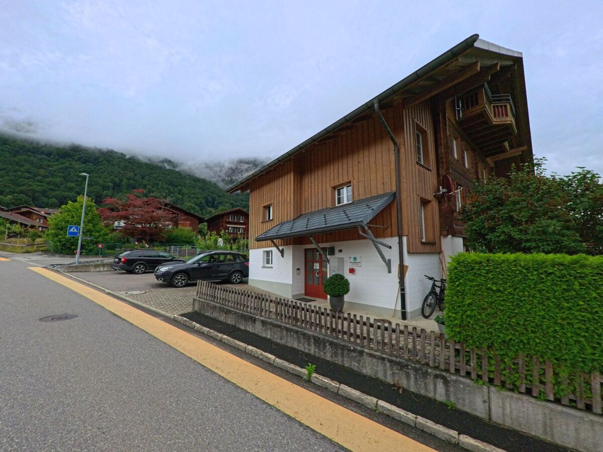 In Brienzwiler (3856, Kanton Bern) steht ein modernes Holzhaus mit Vorgarten und Vordach an einer ruhigen Strasse mit Parkplätzen, umgeben von dichter Vegetation und nebelverhangenen Hügeln.