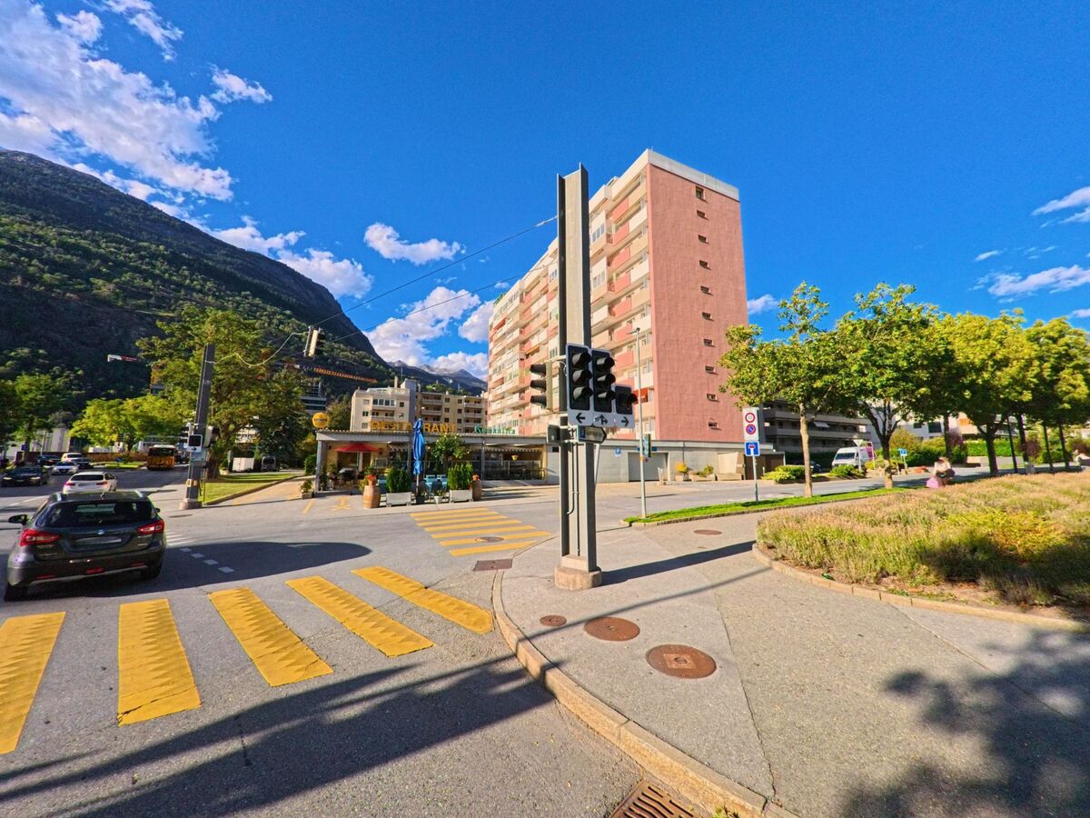 Ein hohes Wohngebaeude mit Balkonen steht in Brig-Glis (3900, Kanton Wallis) an einer belebten Strassenkreuzung mit Fussgaengerstreifen und Ampeln.