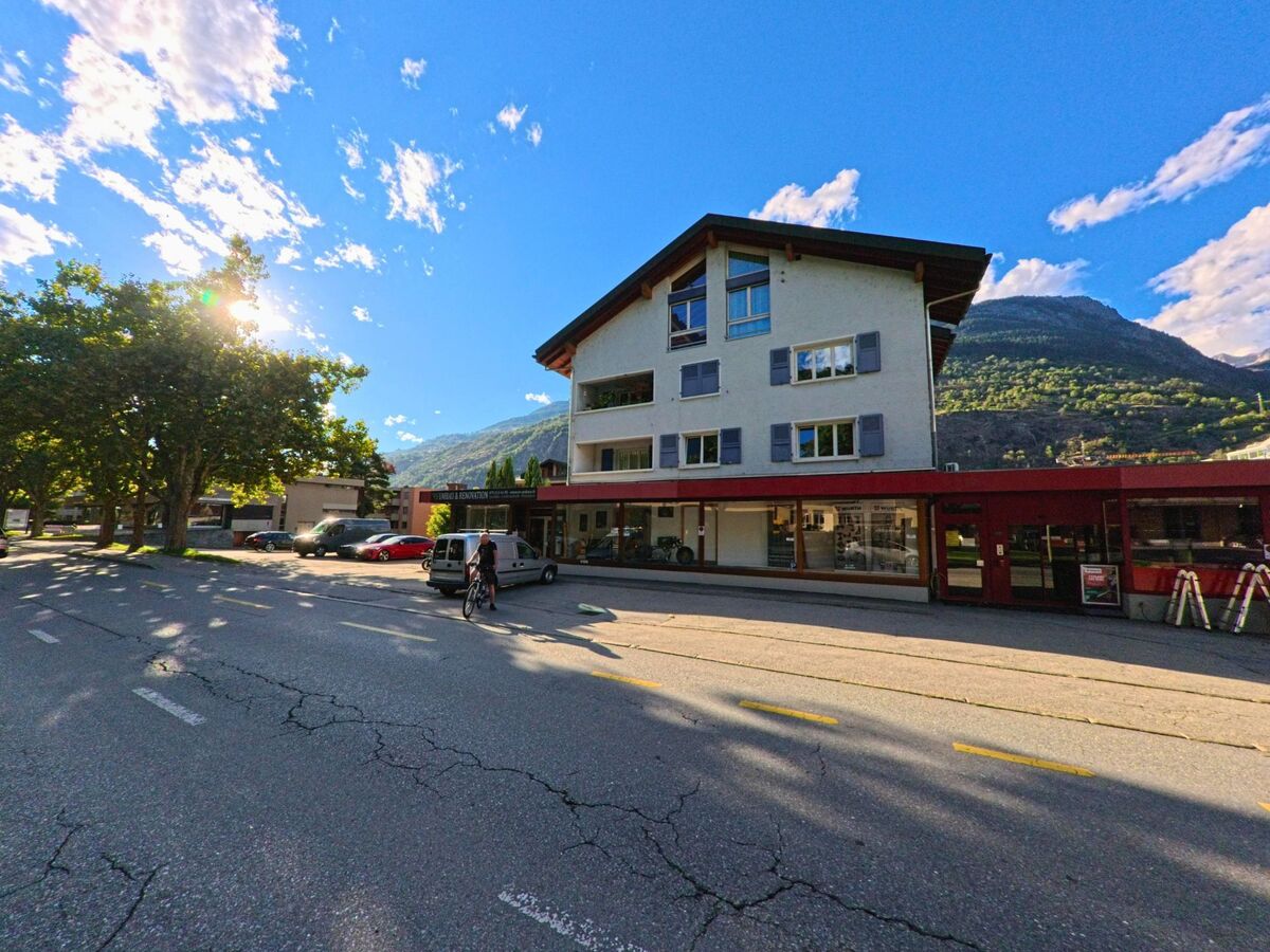 Ein Wohn- und Geschaeftshaus in Brig-Glis (3900, Kanton Wallis) steht an einer Strasse, waehrend im Hintergrund die bewaldeten Berge aufragen.