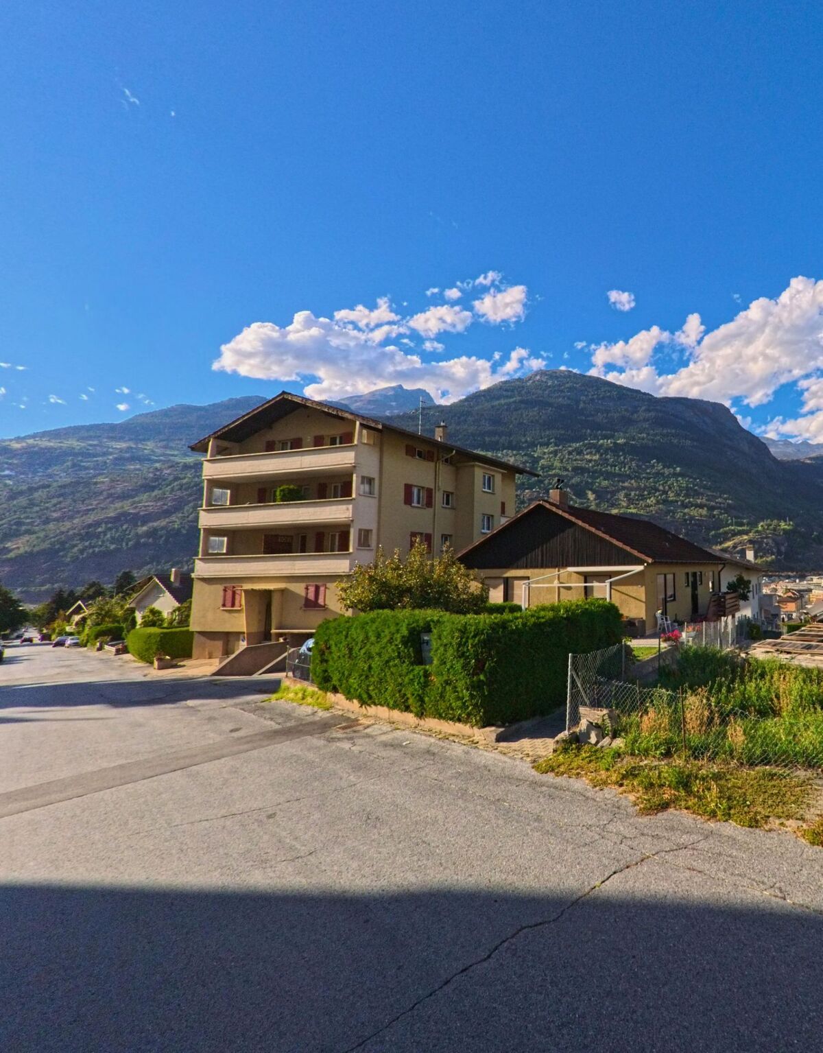 Ein Wohnhaus mit Balkonen steht in Brig-Glis (3900, Kanton Wallis) am Rand einer Quartierstrasse mit Blick auf die umliegenden Berge.