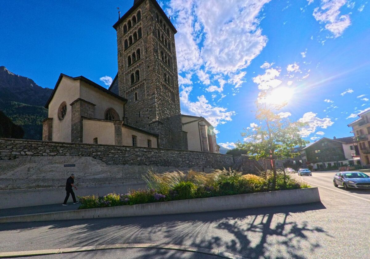 Die Sonne beleuchtet den markanten Kirchturm in Brig-Glis (3902, Kanton Wallis), der sich über einer steinernen Mauer erhebt.